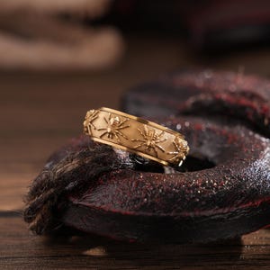May include: A gold-colored ring with a repeating spider design. The ring is set against a dark, textured background, with a wooden surface below. The ring's intricate details are highlighted, showcasing the spider motif.