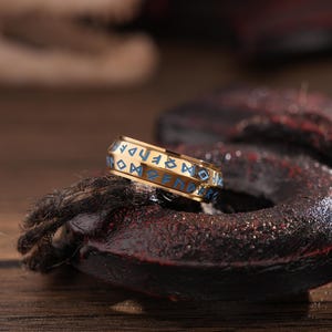May include: A gold ring with blue enamel runes inscribed around the band. The ring is on a dark brown surface.