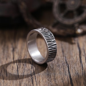 Puede incluir: Un anillo de plata con un diseño texturizado de madera. El anillo está sobre una superficie de madera.