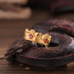 Può includere: Un anello a corona dorata con pietre preziose rosse. L'anello presenta un design dettagliato con croci ed elementi decorativi. L'anello è posto su una superficie scura e strutturata, con sfondo in legno.