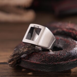 Puede incluir: Un anillo de plata con un espacio abierto de forma cuadrada en el centro. El anillo está sobre una superficie marrón oscura.