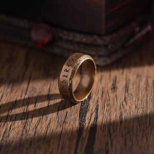 Può includere: Un anello color bronzo con simboli runici incisi. L'anello è posto su una superficie di legno, con uno sfondo scuro e sfocato. Il design dell'anello presenta una fascia di caratteri incisi attorno alla sua circonferenza, suggerendo un tema storico o fantasy.