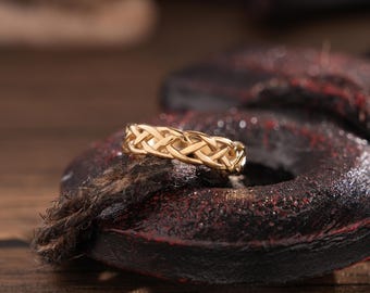 May include: A gold-colored ring with a woven design. The ring is set against a dark, textured background. The ring's intricate pattern is highlighted by the lighting, showcasing its detailed craftsmanship. The ring is a piece of jewelry.
