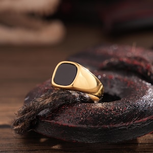 May include: A gold-colored signet ring with a square, black onyx stone. The ring is displayed on a dark, textured surface with a wooden background. The ring's band is polished and reflects light.
