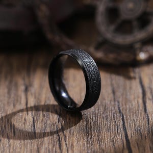 May include: A black ring with a textured surface and an inscription. The ring is made of metal and has a simple design.