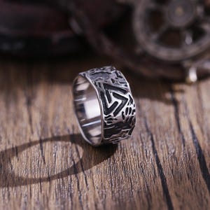 May include: A silver ring with a textured design featuring a triangular symbol. The ring is on a wooden surface.