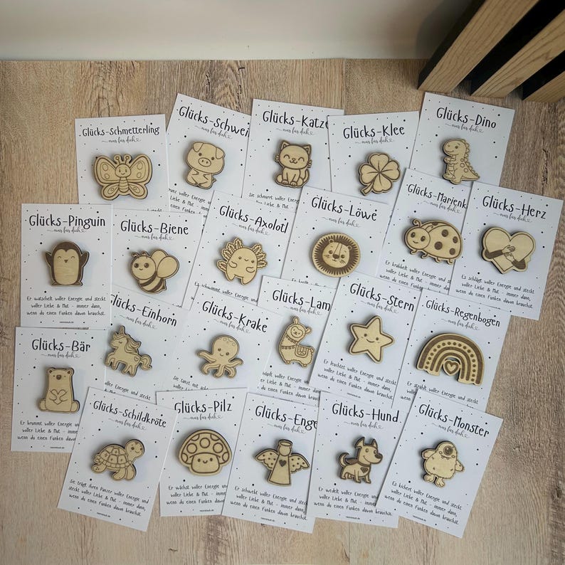 May include: A collection of wooden pins with various designs, including a butterfly, pig, cat, four-leaf clover, dinosaur, ladybug, and heart. Each pin is on a white card with German text. The pins are made of light-colored wood.
