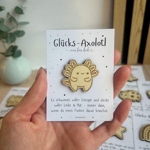 May include: A wooden axolotl pin with the text "Gl&uuml;cks-Axolotl nur f&uuml;r dich" on a white card. The axolotl is light brown with a smiling face. The card also has text in German.