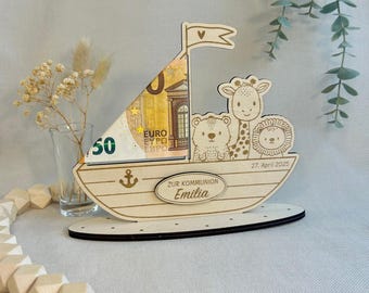 Könnte beinhalten: Holzboot-Sparschwein mit einem 50-Euro-Schein im Inneren. Das Boot ist mit einer Giraffe, einem Löwen und einem Bären verziert. Das Boot trägt den Text "Zur Kommunion Emilia" und "27. April 2025".