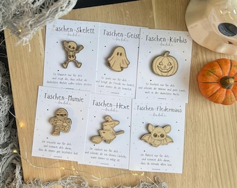May include: A collection of six wooden Halloween-themed pins on cards. The pins feature a skeleton, ghost, pumpkin, mummy, witch on a broom, and bat. Each card has German text. An orange pumpkin is also visible.