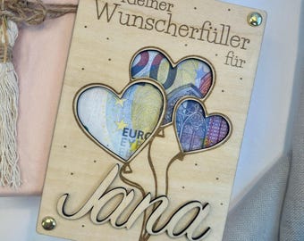 Könnte beinhalten: Holzkärtchen mit dem Text "Kleiner Wunscherschfiller fur" und dem Namen "Jana" in Kursivschrift. Die Karte hat drei herzförmige Ausschnitte, in denen Euro-Scheine zu sehen sind.