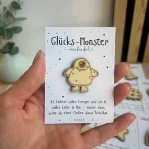 May include: A wooden Gl&uuml;cks-Monster pin, featuring a cartoon monster with one eye, is displayed on a white card. The card has black text and small black dots. The pin is held in a hand.