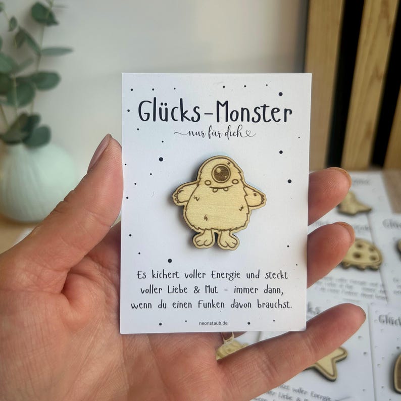 May include: A wooden Gl&uuml;cks-Monster pin, featuring a cartoon monster with one eye, is displayed on a white card. The card has black text and small black dots. The pin is held in a hand.