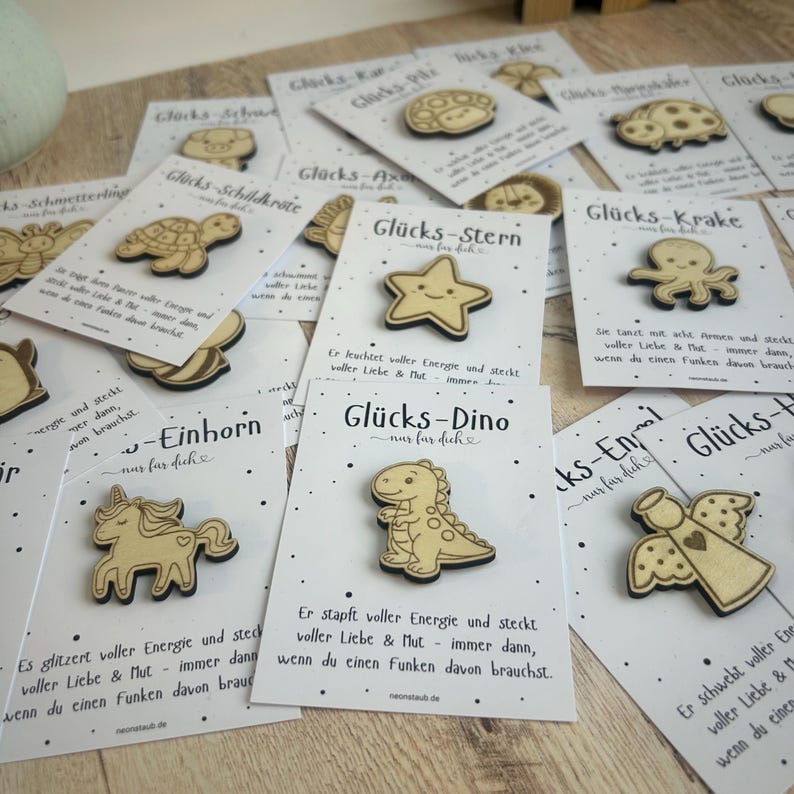 May include: Wooden pins with various designs, including a star, dinosaur, and unicorn, are displayed on white cards. Each pin is laser-cut and features a unique shape. The cards have German text and a dotted pattern.