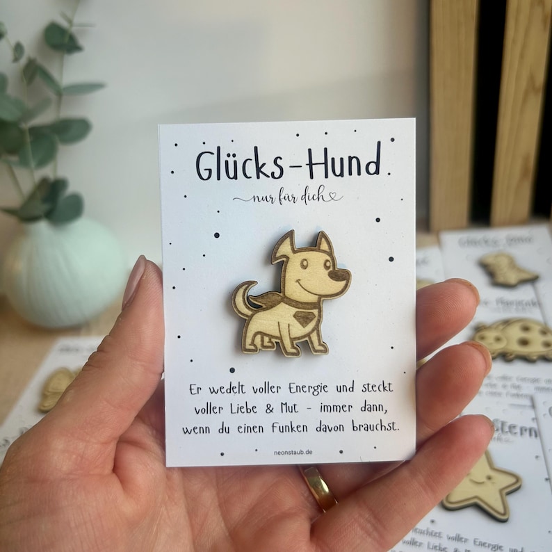 May include: A wooden dog-shaped pin with the German text "Gl&uuml;cks-Hund" on a white card. The pin is light brown with a smiling face and a heart-shaped design. The card has additional German text and small black dots.