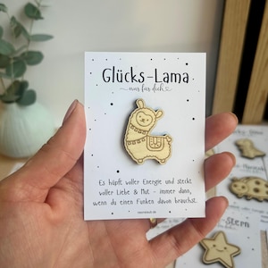 May include: A wooden llama pin on a white card with German text. The pin is light brown with etched details. The card has the words "Gl&uuml;cks-Lama" and "nur f&uuml;r dich" printed on it, along with additional text and a website address.