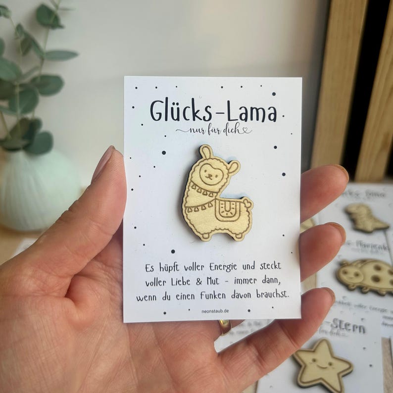 May include: A wooden llama pin on a white card with German text. The pin is light brown with etched details. The card has the words "Gl&uuml;cks-Lama" and "nur f&uuml;r dich" printed on it, along with additional text and a website address.