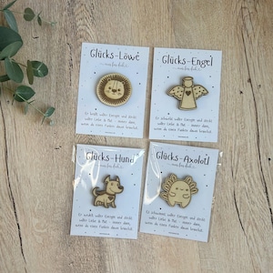 May include: Four wooden pins on cards, each featuring a different design: a lion, an angel, a dog, and an axolotl. The cards have German text and a small illustration of each animal. The pins are light brown.
