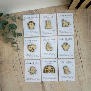 May include: A collection of wooden pins with various designs, including a penguin, bear, unicorn, bee, llama, monster, octopus, rainbow, and dinosaur. Each pin is on a card with German text.