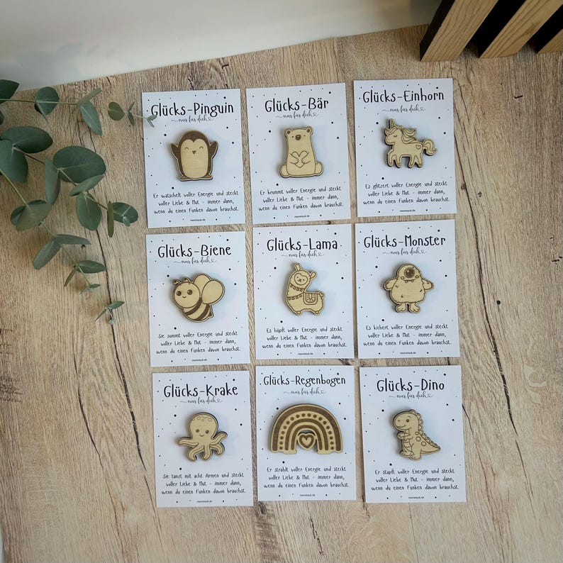 May include: A collection of wooden pins with various designs, including a penguin, bear, unicorn, bee, llama, monster, octopus, rainbow, and dinosaur. Each pin is on a card with German text.