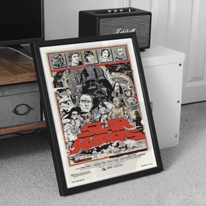May include: Framed Star Wars movie poster featuring a collage of characters and the title in large red letters. The poster has a black frame and is displayed on a carpeted floor.