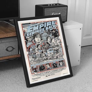 May include: Framed poster featuring artwork from "The Empire Strikes Back" with a black frame. The poster has a cream background with blue and red accents. The artwork includes characters and scenes from the film.