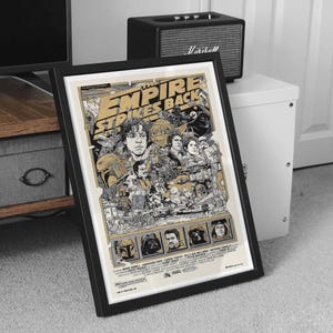 May include: Framed poster for "The Empire Strikes Back" with a black frame. The poster features a detailed illustration with characters and scenes from the film, in shades of beige, black, and gold. The title is prominently displayed at the top.