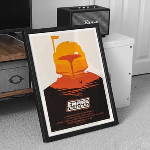 May include: Framed poster featuring a silhouette of Boba Fett's helmet in shades of orange and yellow against a cream background. The poster includes the text "Star Wars: The Empire Strikes Back" and is in a black frame.