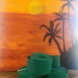 May include: Four green rolled up strips of fabric, possibly for crafts or home decor, sitting on a brown surface in front of a painting of a sunset with palm trees.