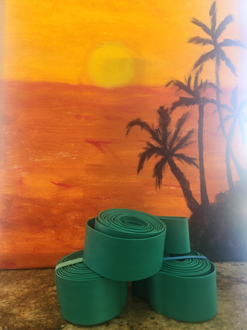 May include: Four green rolled up strips of fabric, possibly for crafts or home decor, sitting on a brown surface in front of a painting of a sunset with palm trees.