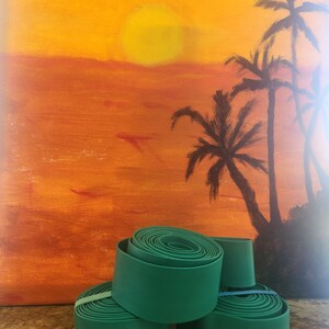 May include: Four rolls of teal green fabric tape stacked on top of each other. The tape is rolled up and has a smooth surface. The background is a painting of a sunset with palm trees.