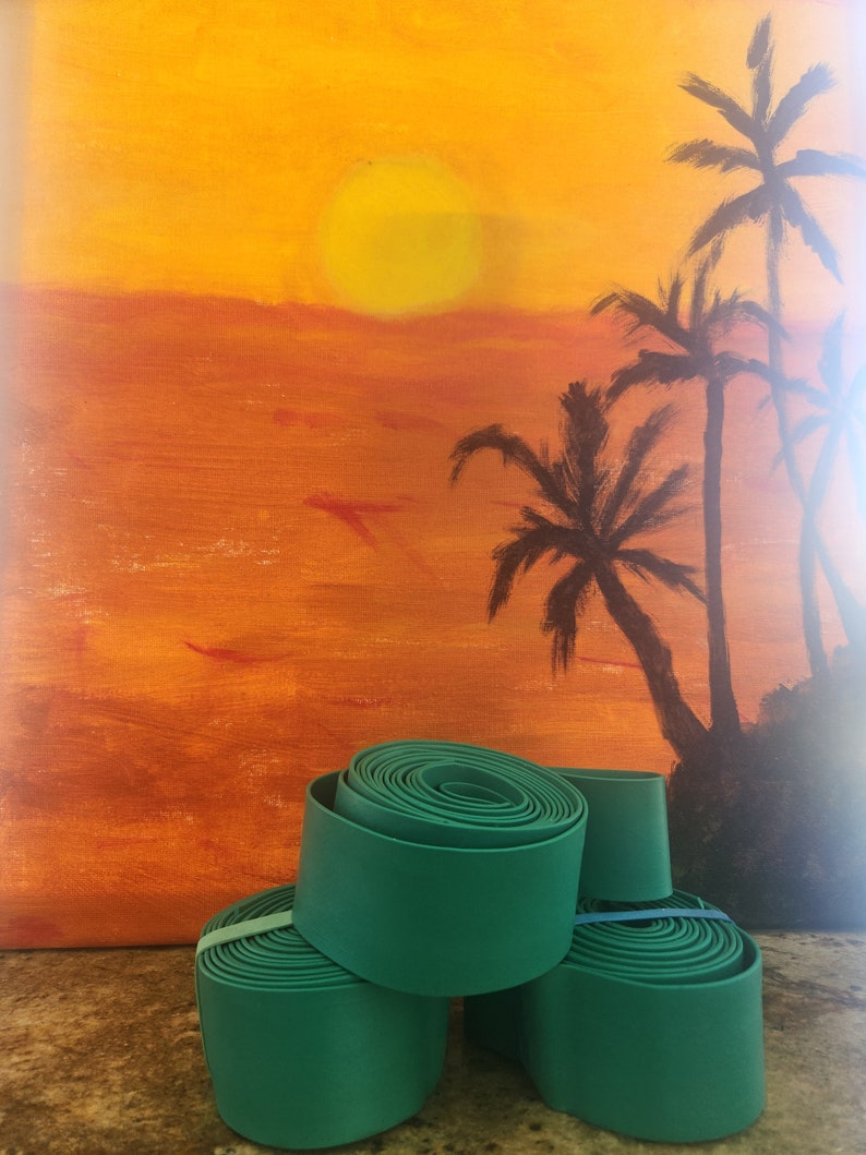 May include: Four rolls of teal green fabric tape stacked on top of each other. The tape is rolled up and has a smooth surface. The background is a painting of a sunset with palm trees.