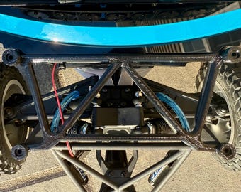 May include: Close-up of a black and gray RC car chassis. The chassis features a complex geometric design with visible suspension components, tires, and a blue accent. The tires have a rugged tread pattern.