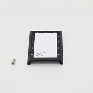 May include: A black rectangular component with a white center panel featuring a stylized 'X' logo. The component has small, evenly spaced dots around the perimeter and a silver screw is next to it. The item is likely a part of a larger device.