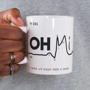 Op de afbeelding: Een witte keramische mok met een zwarte EKG-lijn en de tekst "OH MI" en "I THINK WE MIGHT NEED A CHAIR!" De EKG-lijn heeft labels voor de verschillende delen van de hartslag, waaronder "P", "Q", "R", "S", "T" en "J".