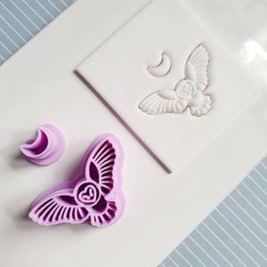 May include: A set of two purple plastic cookie cutters in the shape of an owl with a crescent moon. The cutters are on a white surface with a blue and white striped background.