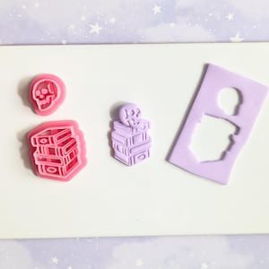 Puede incluir: Una colección de cortadores de arcilla polimérica en rosa y morado. Los cortadores tienen forma de calavera, una pila de libros y una calavera encima de una pila de libros. Un cortador es un espacio negativo de una calavera.