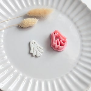 May include: A white ceramic plate with a fluted edge holds a small, speckled white ghost-shaped item and a pink ghost-shaped cookie cutter. Two dried, beige bunny tail grass stems are also on the plate, creating a Halloween theme.