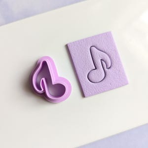 May include: A purple plastic cookie cutter shaped like a musical note, next to a purple fondant imprint of the same note.