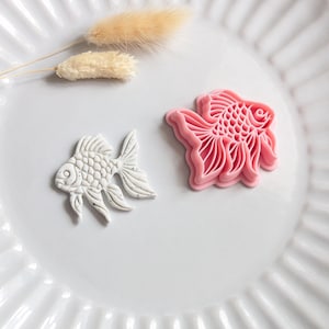 May include: A pink plastic goldfish-shaped cookie cutter alongside a white goldfish-shaped cutout with a speckled texture. The cutter features intricate internal lines for detailing the fins and scales of the fish, while the cutout displays the embossed pattern.