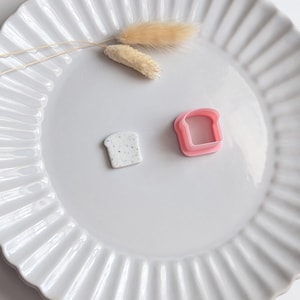 Peut inclure: Une assiette en céramique blanche à bordure cannelée contient un petit objet rectangulaire tacheté et un emporte-pièce rose en forme de pain. Des herbes décoratives séchées de couleur beige sont également sur l'assiette. Les objets sont disposés sur l'assiette.