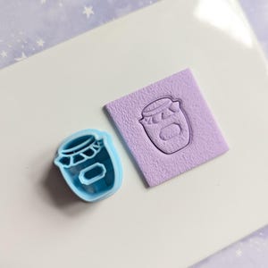 May include: A blue plastic cookie cutter shaped like a jar with a lid and a heart on top. Next to it is a purple rubber stamp with the same design.