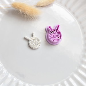 Puede incluir: Dos herramientas artesanales con temática de hilo. Una es una bola de hilo blanca y texturizada con dos pequeños clavijas. La otra es un cortador morado con forma de bola de hilo. Una ramita de hierba beige seca se encuentra encima.