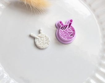 Puede incluir: Dos herramientas artesanales con temática de hilo. Una es una bola de hilo blanca y texturizada con dos pequeños clavijas. La otra es un cortador morado con forma de bola de hilo. Una ramita de hierba beige seca se encuentra encima.