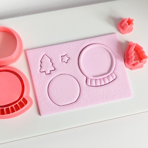 Op de afbeelding: Roze plastic koekjessnijders in de vorm van een sneeuwbol, kerstboom en ster. De sneeuwbolsnijder heeft een ronde basis en een koepel. Extra boom- en stersnijders zijn ook aanwezig.