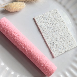 Op de afbeelding: Een roze, getextureerd, cilindrisch object en een wit, rechthoekig object met een gespikkeld patroon worden gepresenteerd op een witte, gegroefde plaat. Gedroogd, beigekleurig decoratief gras is ook aanwezig.