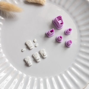 May include: A white ceramic plate displays small, light purple cookie cutters and miniature white and grey coffee cup-shaped decorations. The cutters are in various shapes, including a coffee cup and a lid. A dried, beige plant stem is also on the plate.