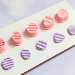 May include: A set of pink and purple polymer clay moulds and shapes. The moulds are designed with a ribbed pattern and include a circle, triangle, and oval shape. The shapes are also ribbed and include a circle, oval, and triangle.