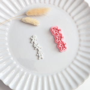 Peut inclure: Un emporte-pièce en plastique rose en forme de pile verticale de trois bonshommes en pain d'épice, accompagné d'une découpe en argile ou en pâte blanche du même motif, avec des détails faciaux et des boutons en relief.