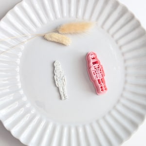 Könnte beinhalten: Ein rosafarbener Nussknacker-Ausstecher aus Kunststoff und ein kleines, weiß gesprenkeltes, nussknackerförmiges Ausstechstück. Die Gegenstände liegen auf einem weißen, geriffelten Teller neben zwei kleinen, hellen, getrockneten Pflanzenteilen.